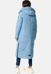 Lang, lichtblauwe pufferjas met een chevron-quiltpatroon, grote capuchon en zijritsen, gecombineerd met donkere jeans en stevige witte sneakers.