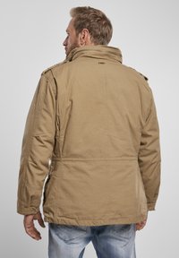 Jachetă khaki cu guler înălțat, epoleți pe umeri, buzunare laterale și detalii cusute contrastante. Materialul pare texturat și durabil.
