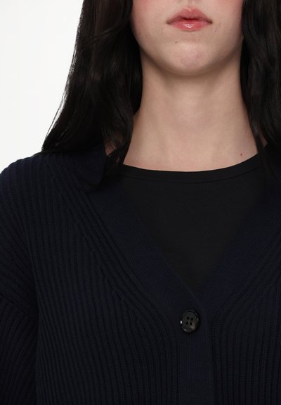Marineblauer geripptes Strickjacke mit einem tiefen V-Ausschnitt und einem einzelnen schwarzen Knopfverschluss, über einem schwarzen Oberteil geschichtet, das ein strukturiertes Strickdesign aufweist.