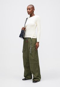 Hosszú ujjú, krém színű felső oldalhasítékokkal, laza olívazöld cargo nadrággal, amely zsebekkel és színes húzózsinórokkal rendelkezik, valamint fekete szandállal.