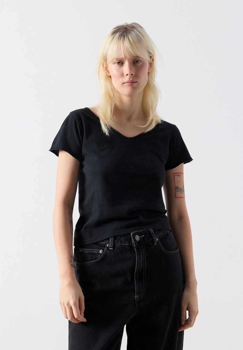 Plavolaska ženska s ravnimi lasmi, oblečena v črno majico z V-izrezom in črne kavbojke, stoji pred enobarvnim belim ozadjem.