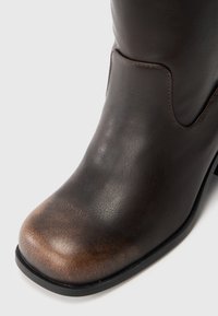Botim de tornozelo de couro castanho-escuro com bico redondo, rugas subtis e sola preta sobre um fundo branco.