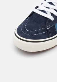 Marineblå semsket sneakers med hvite snører, kontrasterende teal-detaljer, sydd dekorasjon og en kremfarvet gummisåle med teksturert grep.