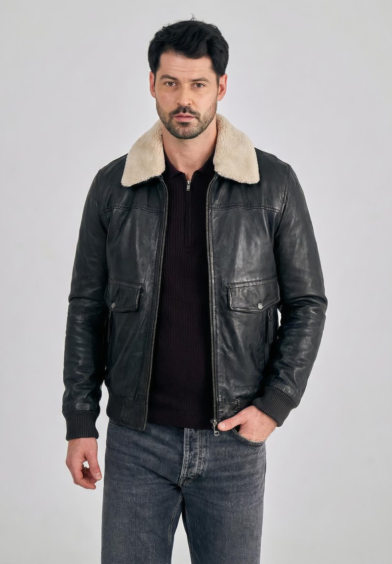 Muž s tmavými vlasy, oblečený v černé kožené bundě se světlým kožíškovým límcem, černém svetru a šedých džínách, stojící před jednobarevným pozadím.