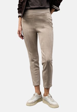 Persoon draagt beige slim-fit broek met voorste naden en elastische tailleband, gecombineerd met witte sneakers en een donker topje.