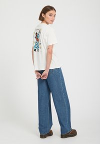 Femme debout, dos tourné, portant un t-shirt blanc avec un motif coloré et un jean bleu à jambes larges avec des chaussures marron sur un fond uni.