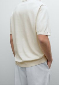 Polo manches courtes couleur crème, texture tricotée, poignets et ourlet côtelés, associé à un pantalon gris clair.