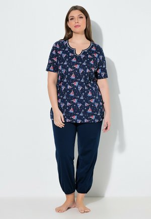 Frau trägt ein marineblaues Kurzarmoberteil mit rotem und weißem Segelbootmuster und passende marineblaue Hosen mit elastischem Bund, steht barfuß.