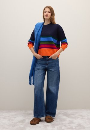 Gekleurde gestreepte trui met marineblauwe, oranje, groene en blauwe tinten, gecombineerd met wijde denim jeans en een blauwe sjaal die over de schouder is gedrapeerd.