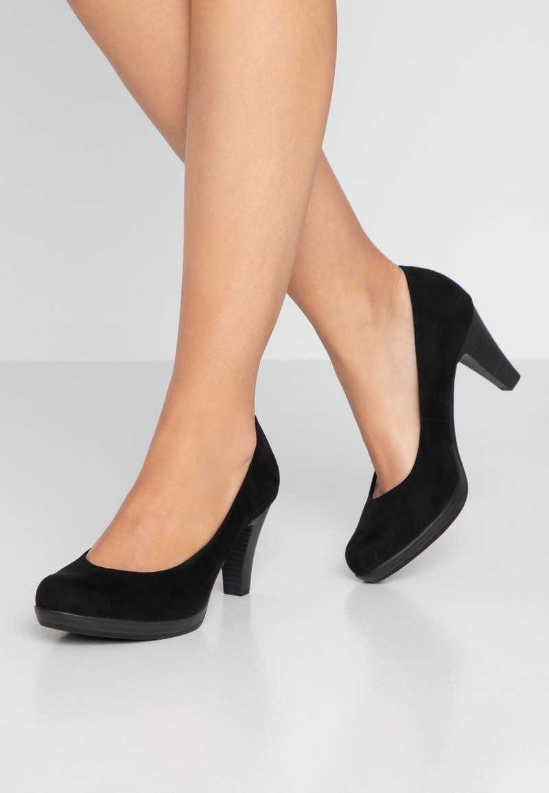 Chaussures à talons hauts en daim noir avec un bout arrondi, une hauteur modeste et un talon bloc texturé, posées sur une surface blanche réfléchissante.