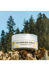 Bocal en plastique transparent avec un couvercle blanc. L'étiquette présente des motifs d'oiseau jaunes et le texte : "SUSANNE KAUFMANN Beurre Corporel pour les Sens."