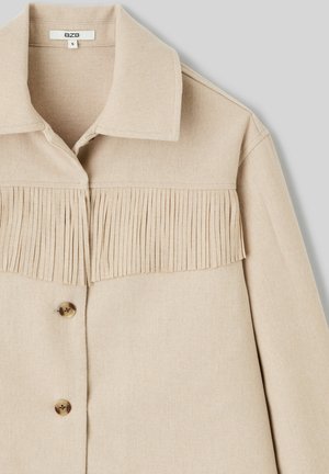 Veste beige avec un col à revers, présentant un design à franges sur la poitrine et deux boutons dorés à l'avant. Confectionnée en tissu texturé.