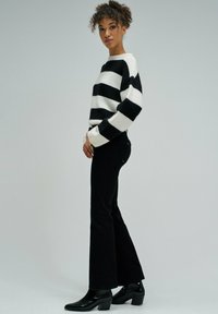 Mujer de pie con pantalones de campana negros, suéter a rayas blanco y negro, y botas negras de tobillo contra un fondo gris liso.