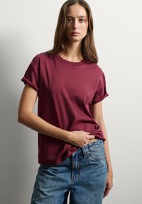 T-shirt in cotone bordeaux con maniche corte arrotolate, scollo rotondo e vestibilità rilassata. Abbinato a jeans in denim blu, per un look casual.