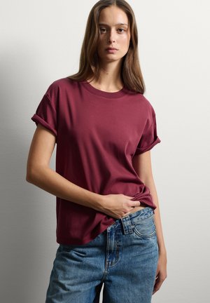 T-shirt in cotone bordeaux con maniche corte arrotolate, scollo rotondo e vestibilità rilassata. Abbinato a jeans in denim blu, per un look casual.