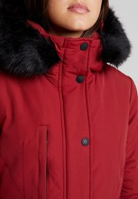 Femme portant un manteau d'hiver rouge avec une capuche bordée de fourrure noire et des boutons-pression noirs, visible des épaules jusqu'aux lèvres sur un fond uni.