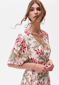 Frau in einem blumenbedruckten V-Ausschnitt-Kleid mit halbenÄrmeln, großen Creolen und lockigen Haaren, die sanft über ihr Gesicht wehen.