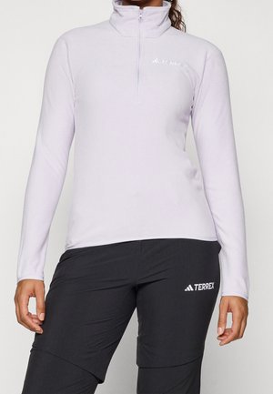 Persoon draagt een lichtlavendelkleurige zip-up top met lange mouwen en zwarte broek, beide met witte Adidas Terrex-logo's.