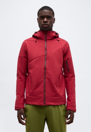 Άνδρας με κόκκινο φούτερ με φερμουάρ και κουκούλα με κορδόνια, φοράει χακί παντελόνι, κοιτάζει προς τα εμπρός με φόντο λευκό.