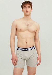 Sive bokser hlače s črnim pasom z blagovno znamko in napisom "JACK & JONES" v belem, ki jih nosi model na nevtralni podlagi.