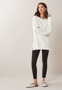 Sudadera blanca de gran tamaño con mangas largas, aberturas laterales, combinada con leggings negros y zuecos crema con un acento dorado.