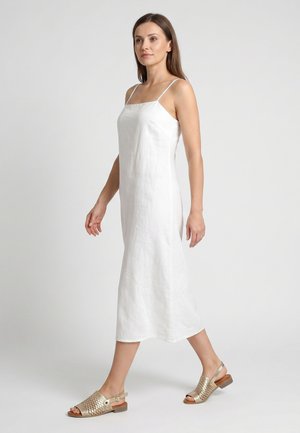Frau geht, trägt ein ärmelloses weißes Midikleid und goldene gewebte Sandalen mit offenem Zeh und niedrigem Absatz vor einem schlichten hellen Hintergrund.
