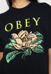 Černé bavlněné tričko s velkým květinovým grafikou v růžové a zelené barvě se slovem "OBEY" v jasně žluté barvě nad designem.