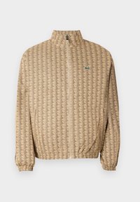 Beige zip-up jakke med et geometrisk mønster. Har en stående krave, elastiske ærmer og et lille logo på brystet.