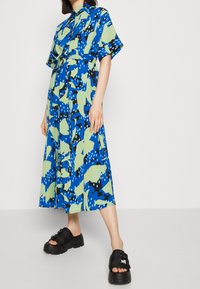 Blau- und grün gemustertes Kleid mit kurzen Ärmeln und einem Bindegürtel; schwarze Plateausandalen mit Schnallen, die kräftige Formen und Farben zur Schau stellen.