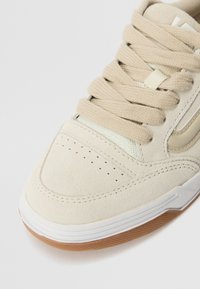 Beige suede sneaker with beige laces, perforated toe box, and white rubber sole. Features a textured side panel with a subtle accent.