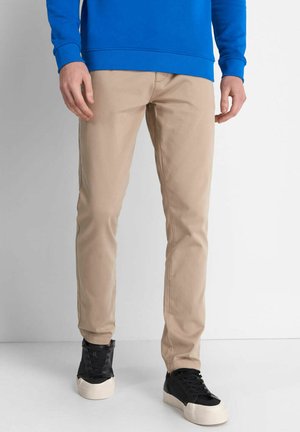 Pantalones de algodón beige claro con un corte ajustado, combinados con zapatillas negras de caña alta que cuentan con una suela blanca gruesa, sobre un fondo neutro.