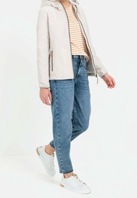 Personne portant une veste blanche zippée, un t-shirt rayé orange et blanc, un jean bleu et des baskets blanches, debout sur un fond blanc.
