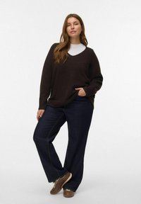 Vrouw staat, gekleed in een donkerbruine V-halstrui over een witte top, donkere wijde spijkerbroek en bruine sneakers tegen een effen achtergrond.
