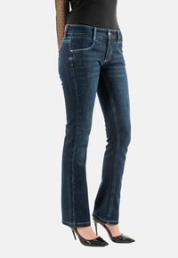 Vaqueros acampanados de mezclilla azul oscuro con un diseño clásico de cinco bolsillos, detalles de costura sutiles y una cintura de altura media. Combinados con tacones negros.
