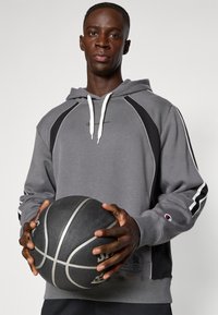 Grå sweatshirt med huva, svarta accenter, vita dragsnören och ribbade manschetter. Håller i en texturerad basketboll. Enfärgad bakgrund.