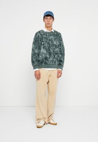 Zaļgans marmora motīvs džemperis ar brīvu piegriezumu, apaļu kaklu un ribinātām piedurknēm, kopā ar krēmkrāsas platajām biksēm un bēša krāsas sporta apaviem.