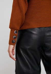 Ein rostfarbener gerippter Pullover mit drei schwarzen Knöpfen an den Bündchen, kombiniert mit schwarzen Lederhosen. Strukturiertes Gewebe und glatte Lederoberfläche.