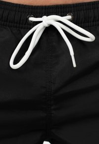 I pantaloni shorts neri presentano un elastico in vita con un cordino bianco. Occhielli metallici sono presenti su entrambi i lati del cordino.