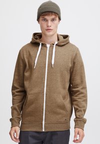 Bruine rits-hoodie met een verstelbare capuchon; heeft een witte rits, zakken en geribbelde manchetten. Draag met een grijze gebreide beanie.