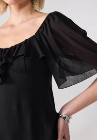 Černé šaty s krátkými průhlednými rukávy, volánkovým výstřihem a hladkým povrchem. Na zápěstí je stříbrný náramek.