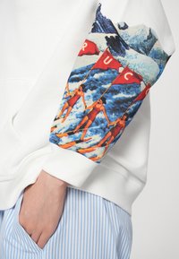Hanorac alb cu un grafic colorat care înfățișează femei în costume de baie și steaguri roșii pe mâneca stângă, asortat cu pantaloni în dungi albastre.