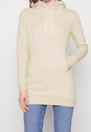 Sweat à capuche beige à manches longues avec cordons blancs et poche avant, porté avec un jean bleu, main reposant dans la poche.