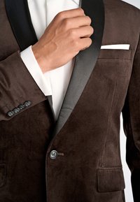 Mann passt den Revers eines braunen Samt-Tuxedo-Jackets mit schwarzem Satin-Kragen und weißem Hemd darunter an.