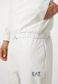 O casaco e as calças de atletismo brancos apresentam um padrão de grelha texturizado, punhos e cintura elásticos, com o logo EA7 bordado nas calças.