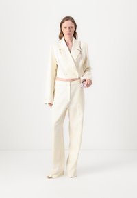 Blazer blanco recortado con botones dobles y pantalones de pierna ancha, con un patrón texturizado. Se sostiene un pequeño bolso rosa en una mano.