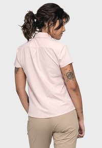 Short-sleeve, light pink shirt made from a smooth fabric with a pointed collar and back pleat detailing, worn with beige pants.