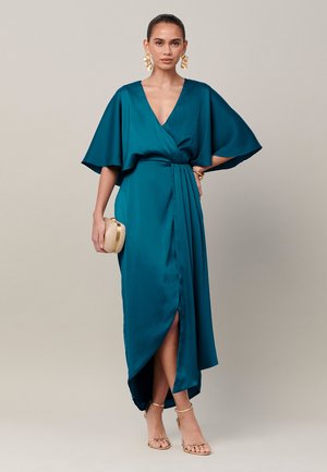 Woman wearing teal wrap dress with wide sleeves, holding a beige clutch, accessorized with large gold earrings and gold sandals.