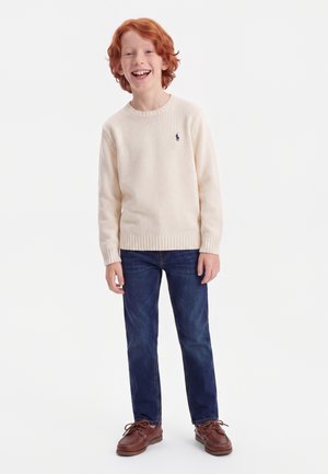 Smilende rødhåret dreng står iført en cremefarvet sweater, mørkeblå jeans og brune snørestøvler mod en hvid baggrund.