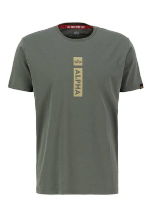 Krátky rukáv olivovo-zeleného trička vyrobeného z bavlny, s vertikálnou zlatou grafikou "ALPHA" na strede a červeným označením "REMOVE BEFORE FLIGHT" vo vnútri goliera.