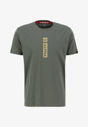 Krátky rukáv olivovo-zeleného trička vyrobeného z bavlny, s vertikálnou zlatou grafikou "ALPHA" na strede a červeným označením "REMOVE BEFORE FLIGHT" vo vnútri goliera.